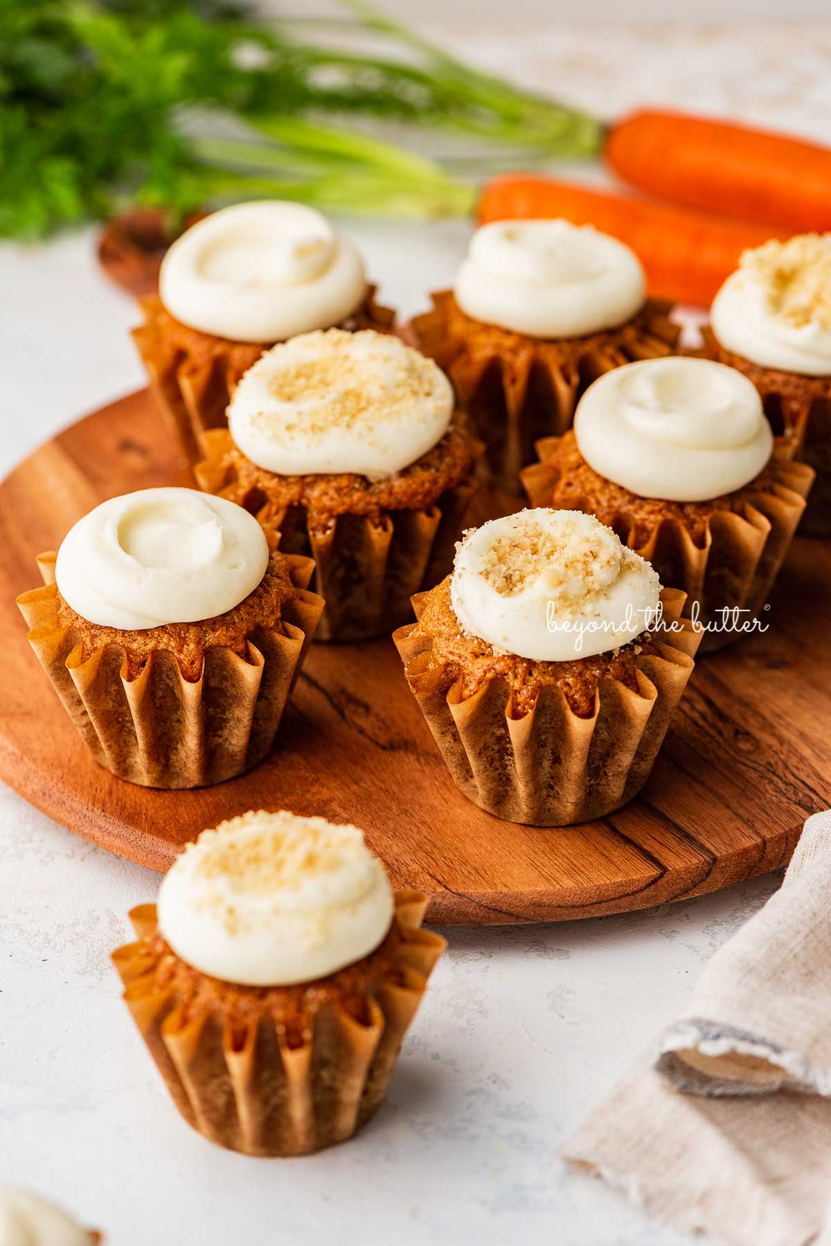 Super moist carrot cake cupcakes with cream cheese frosting placed on a wood cake stand on a white background with some carrots | © Beyond the Butter®