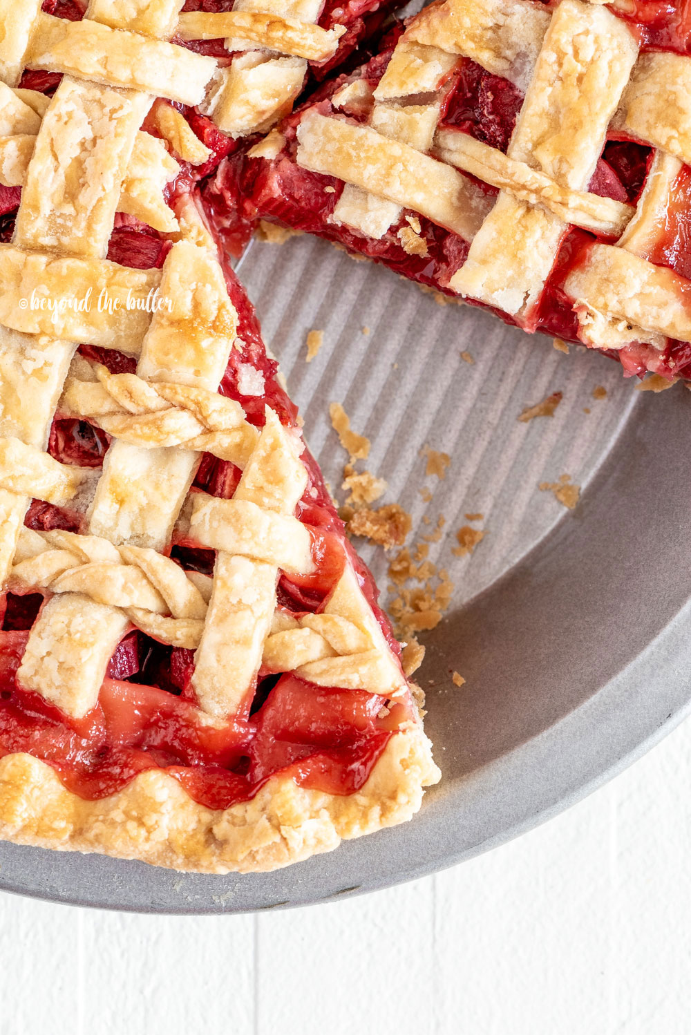 Overhead image of strawberry rhubarb pie with slice removed on white wood table.