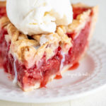 Slice of strawberry rhubarb pie with scoop of vanilla ice cream on dessert plate on white wood table.