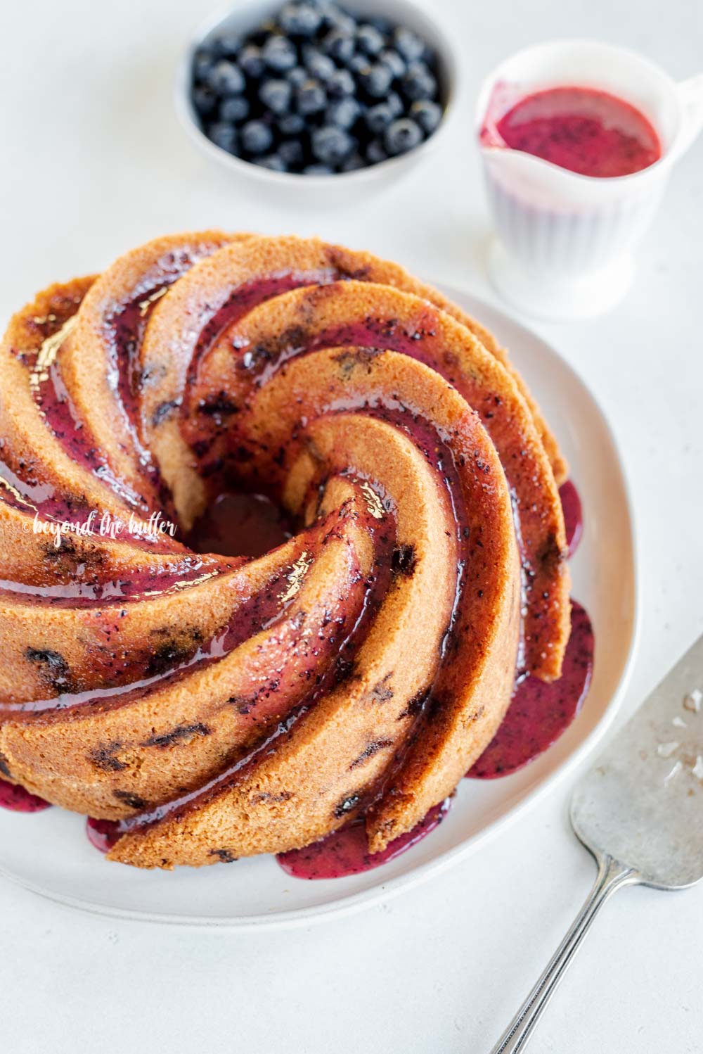 Angled image of super easy blueberry bundt cake with blueberry glaze drizzled over the top | All Images © Beyond the Butter, LLC