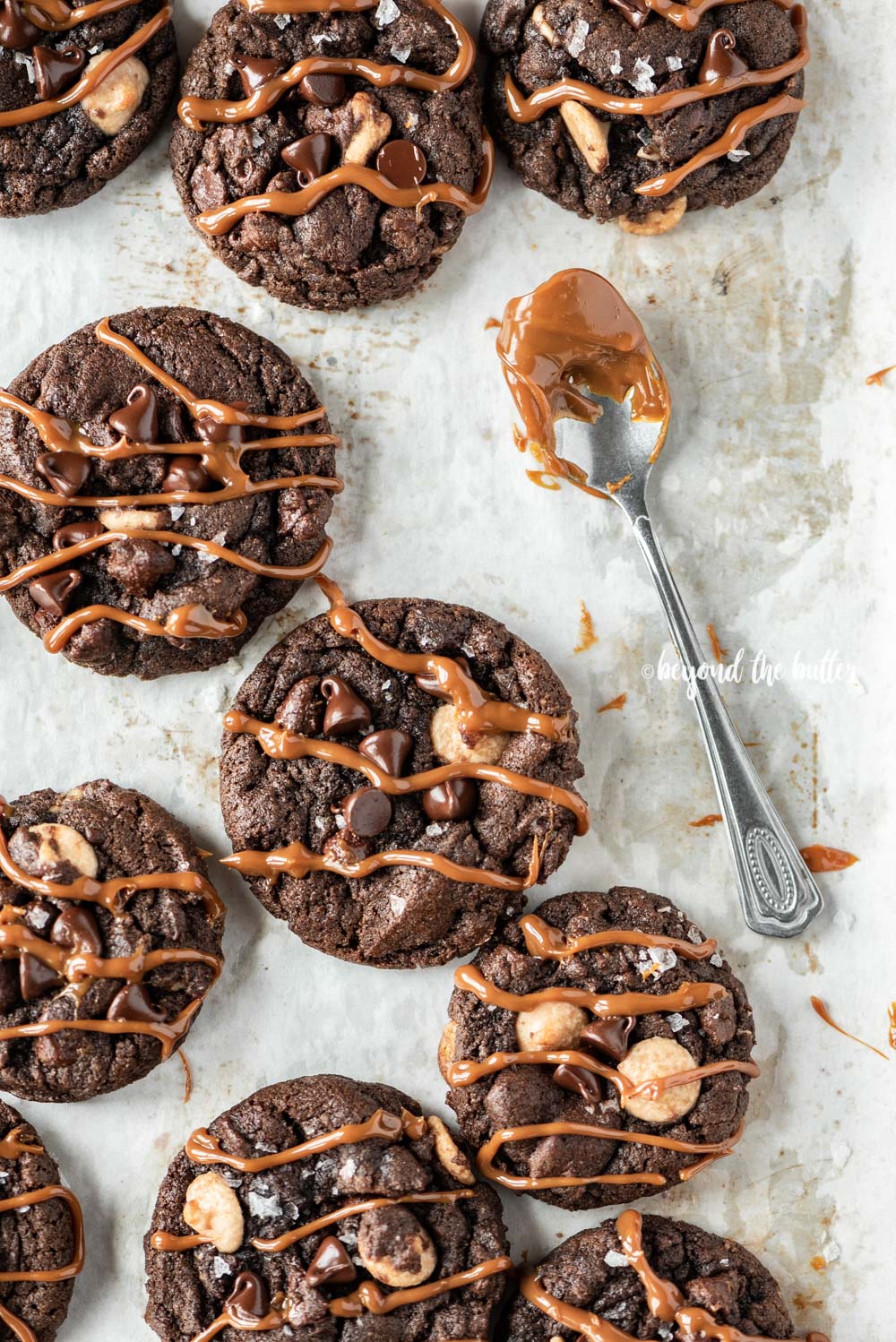Overhead image of just baked double chocolate salted caramel cookies on a baking sheet with spoon dipped in a caramel sauce resting close by | All Images © Beyond the Butter™