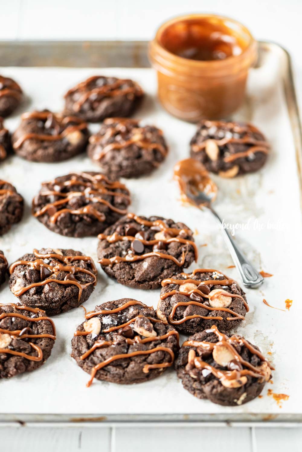Overhead image of just baked double chocolate salted caramel cookies on a baking sheet with a spoon and jar of a caramel sauce resting close by | All Images © Beyond the Butter™