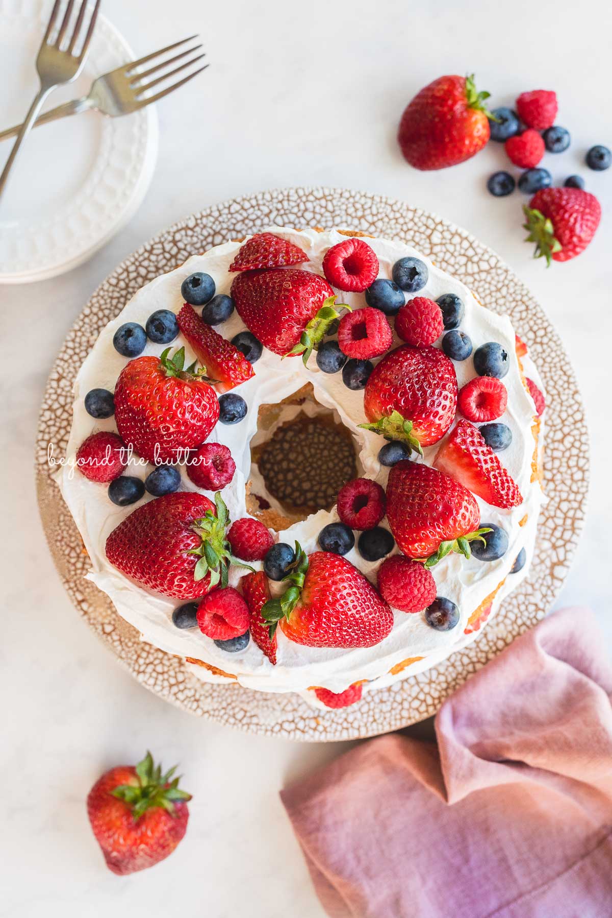 Homemade angel food cake on ceramic cake stand surrounded by dessert plates, forks, berries, and mauve colored cloth napkin on white marbled background | © Beyond the Butter®