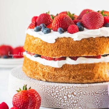 Homemade angel food cake layered with cool whip and fresh berries on a ceramic cake stand with a white background | © Beyond the Butter®