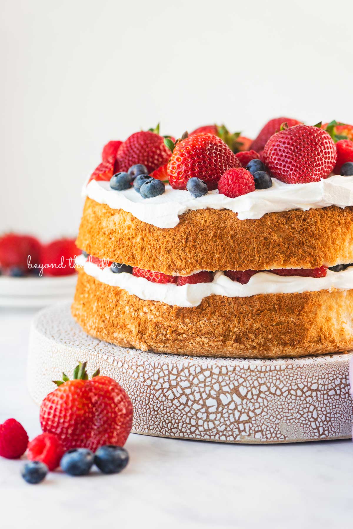 Homemade angel food cake layered with cool whip and fresh berries on a ceramic cake stand with a white background | © Beyond the Butter®