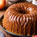 Homemade apple bundt cake with brown sugar glaze surrounded by an apple and chef's knife on dark wood background.