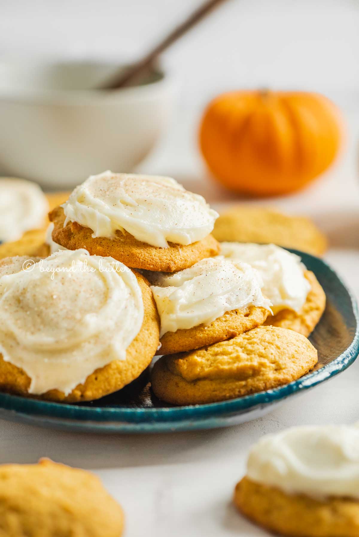 Pumpkin cinnamon cookies topped with cream cheese frosting served on plate with more cookies and pumpkin in the background | All Images © Beyond the Butter™