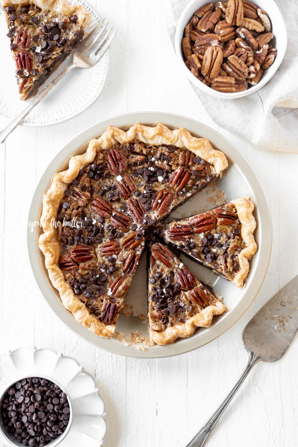 Overhead image showing slices of chocolate pecan pie in pie plate with slices of pie on dessert plates and bowl of chocolate chips and pecans on white wood table.