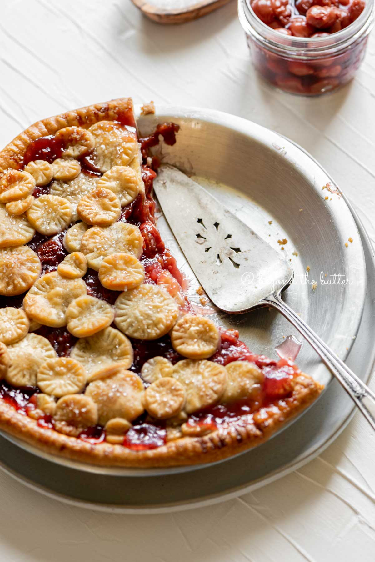 Angled image of half eaten cherry pie with pie server placed on the pie plate | | All Images © Beyond the Butter™