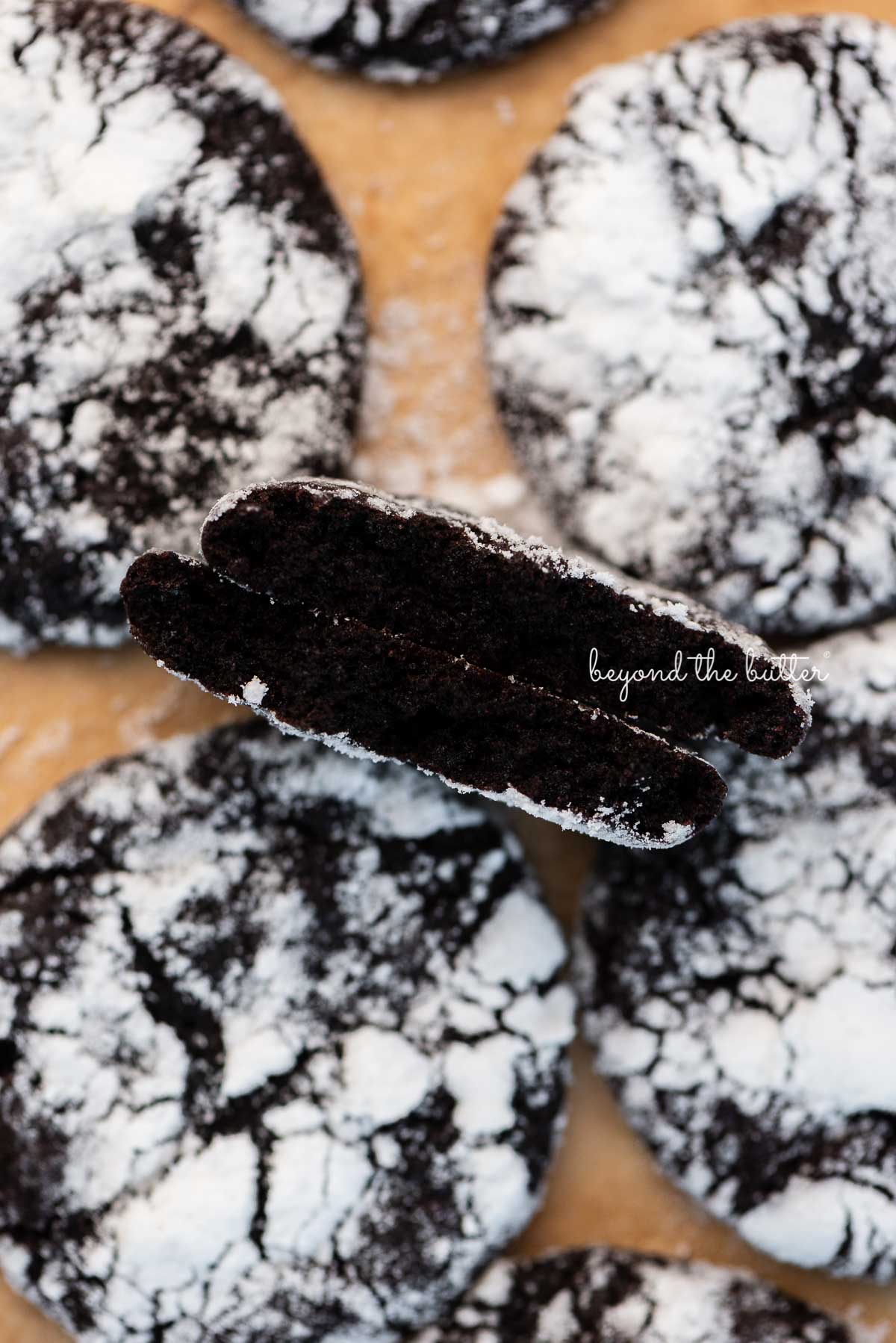 Split dark chocolate crinkle cookie to show the fudgy inside on parchment paper lined baking sheet | © Beyond the Butter®