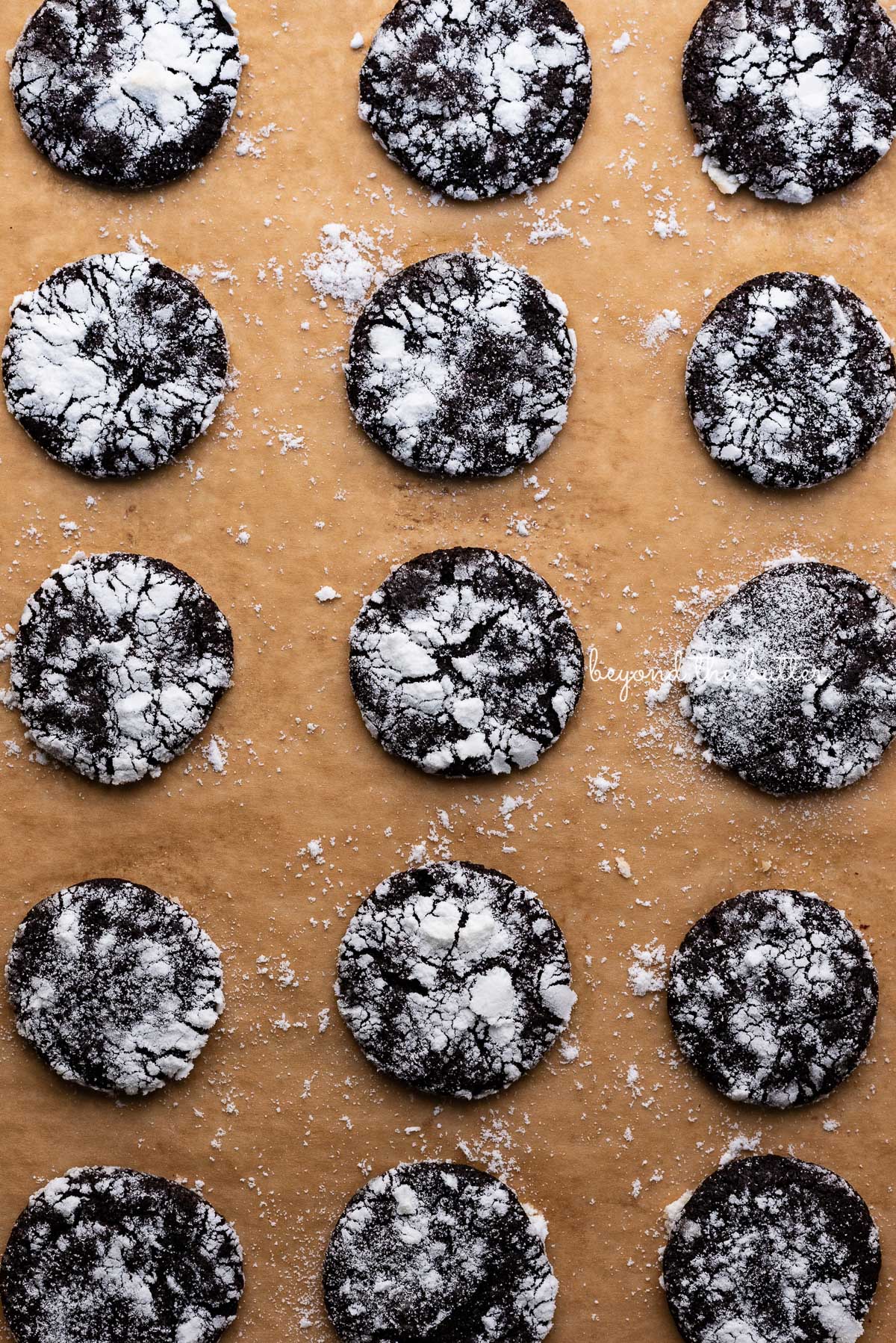 Dark chocolate crinkle cookies evenly spaced on a brown parchment paper lined baking sheet | © Beyond the Butter®