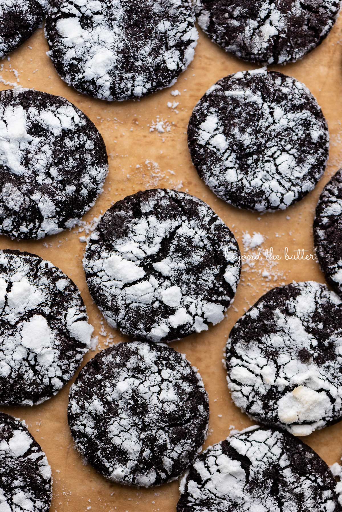 Just baked dark chocolate crinkle cookies on a brown parchment paper lined baking sheet | © Beyond the Butter®