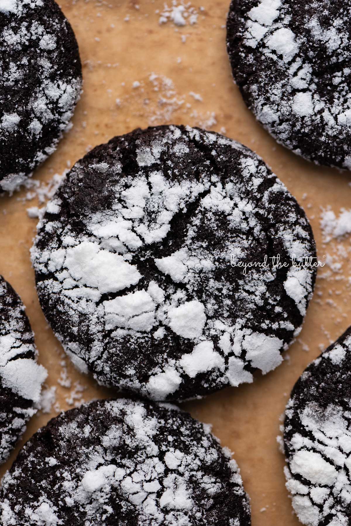 Closeup of dark chocolate crinkle cookie on a brown parchment paper lined baking sheet | © Beyond the Butter®