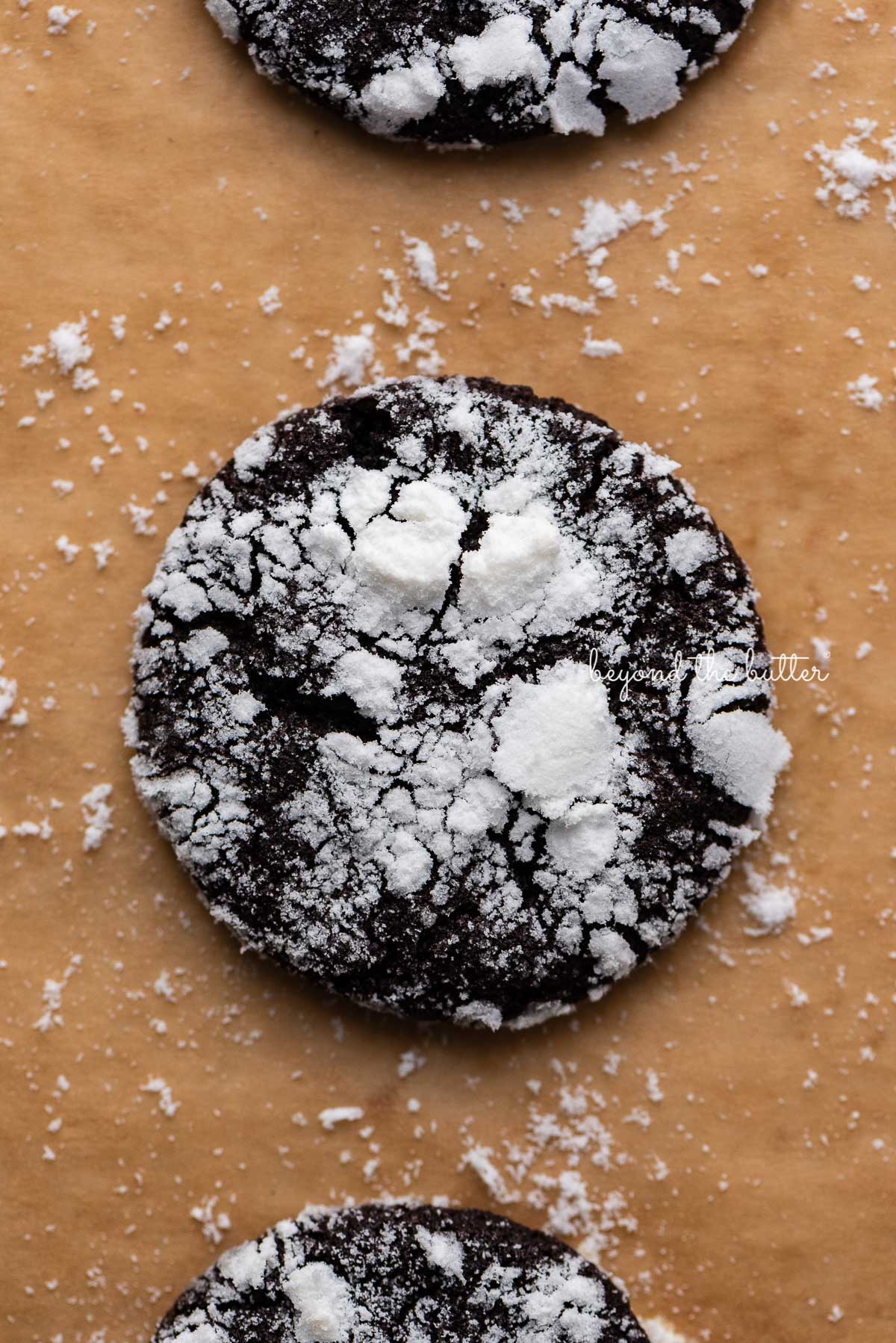 Closeup of dark chocolate crinkle cookie on a brown parchment paper lined baking sheet | © Beyond the Butter®