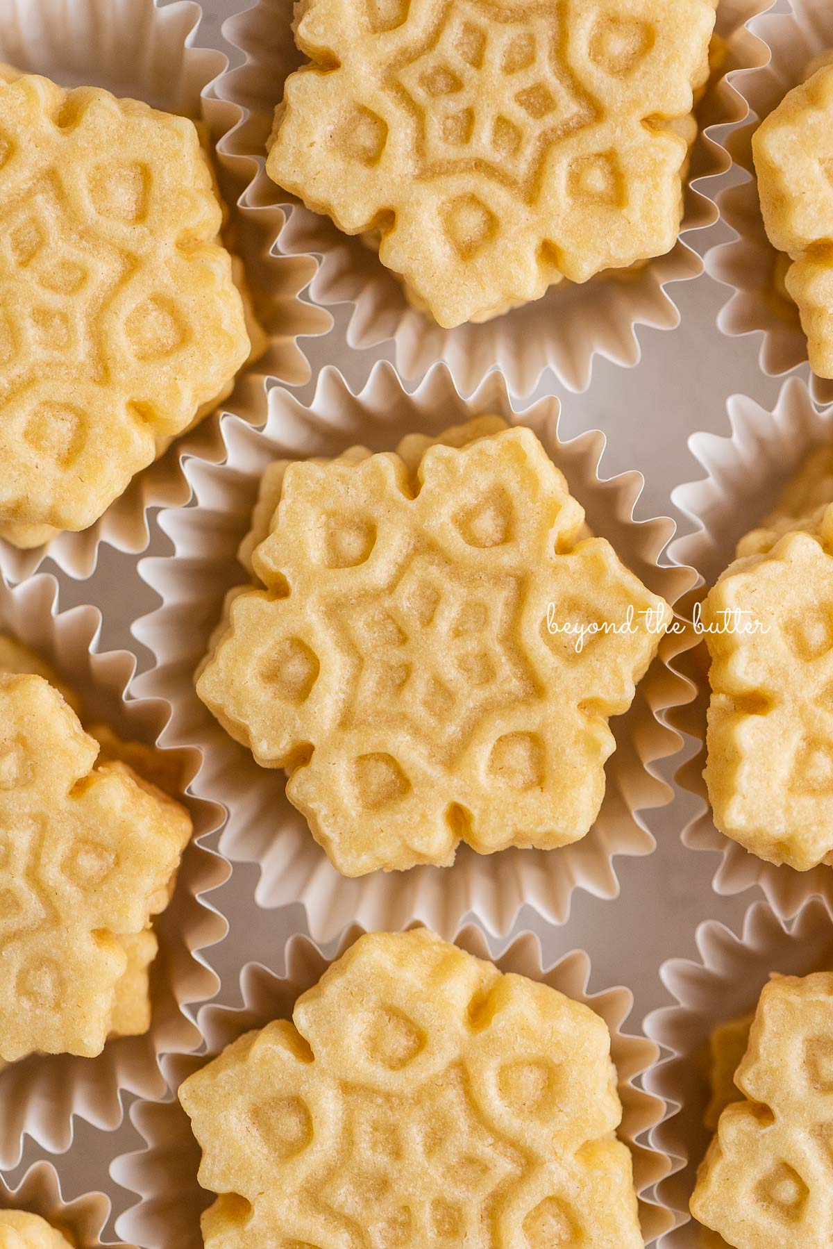Stacks of homemade butter cookies shaped like snowflakes in white cupcake liners | © Beyond the Butter®