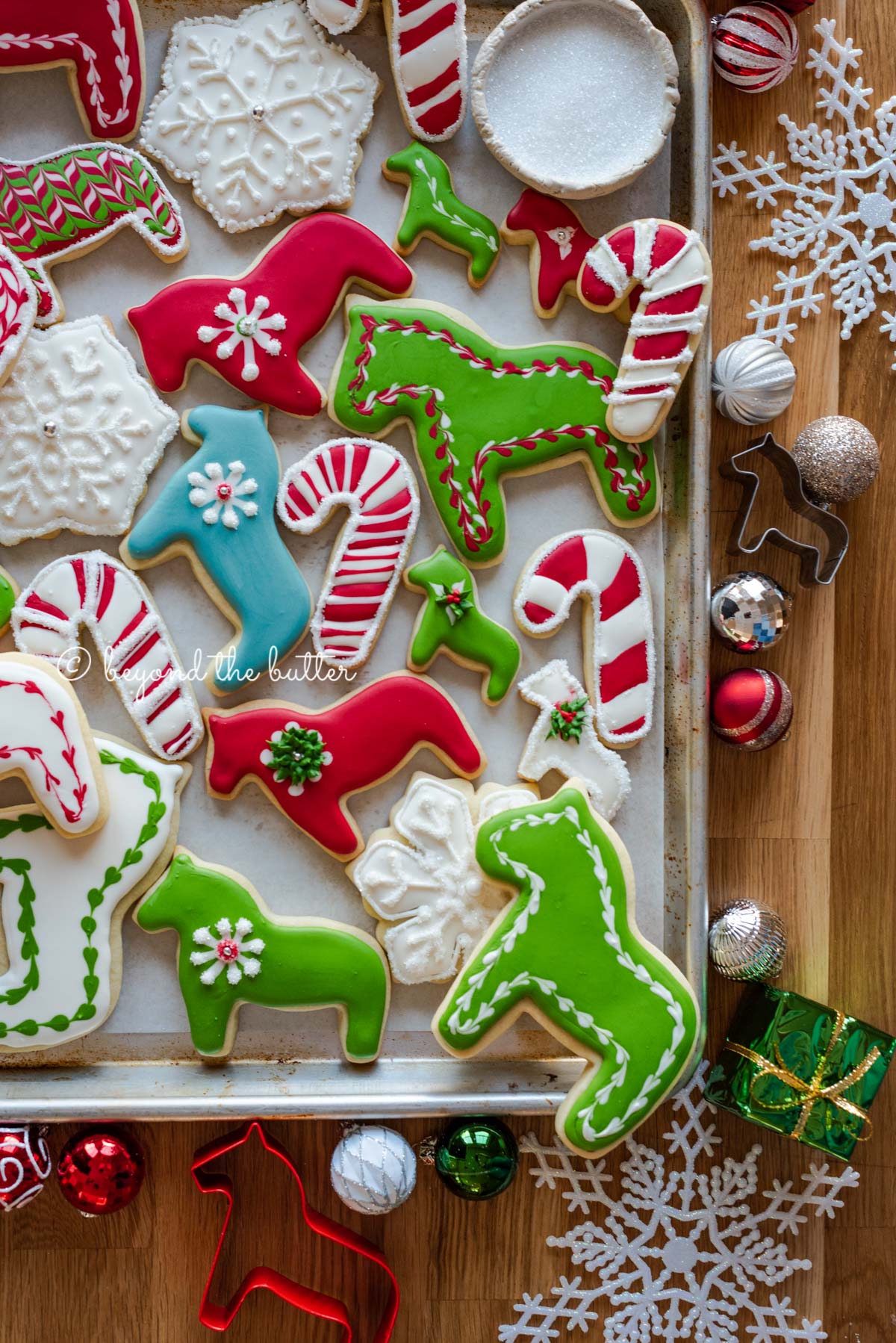 Randomly placed decorated cut out sugar cookies on a parchment lined baking sheet with ornaments and cookie cutters around it | All Images © Beyond the Butter®