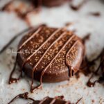 Closeup of peppermint patties on a parchment lined baking sheet sprinkled with cocoa powder and drizzled with melted chocolate.