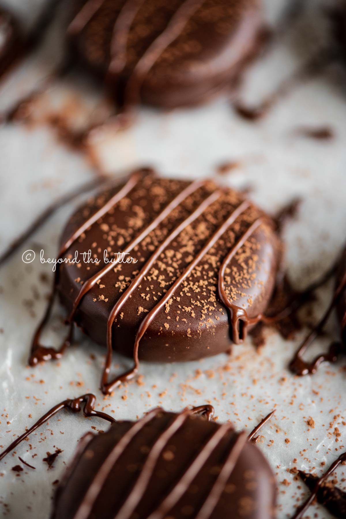 Closeup of homemade peppermint patties on a parchment lined baking sheet sprinkled with cocoa and drizzled with melted chocolate | All Images © Beyond the Butter®