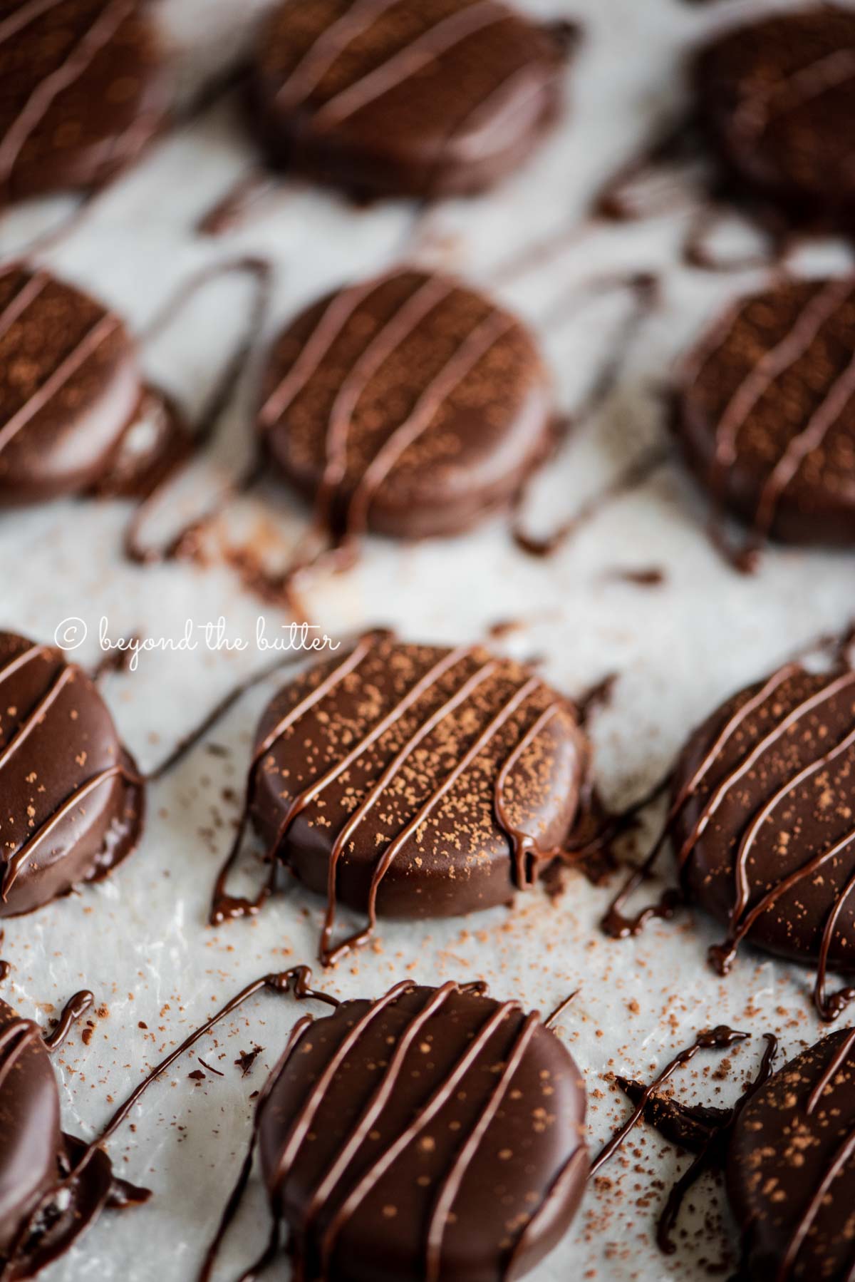 Homemade peppermint patties on a parchment lined baking sheet sprinkled with cocoa and drizzled with melted chocolate | All Images © Beyond the Butter®