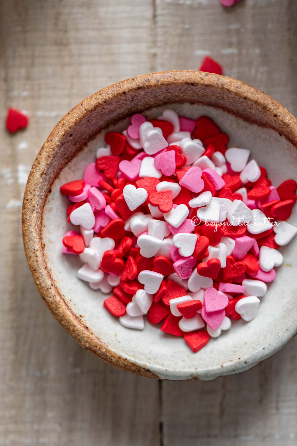 Small bowl of Valentine's Day heart sprinkles Homemade red velvet cupcakes with one opened and half eaten with a small bowl of Valentine's Day sprinkles on light wood background | All images © Beyond the Butter®
