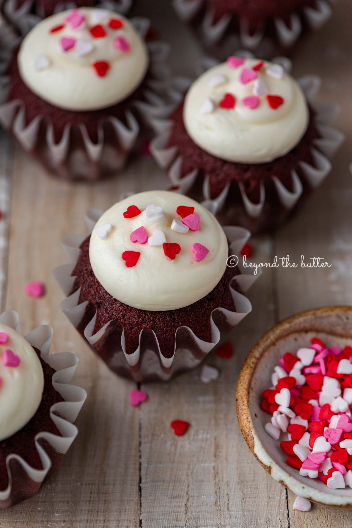 Homemade red velvet cupcakes with cream cheese frosting topped with heart sprinkles on a light wood background | All images © Beyond the Butter®