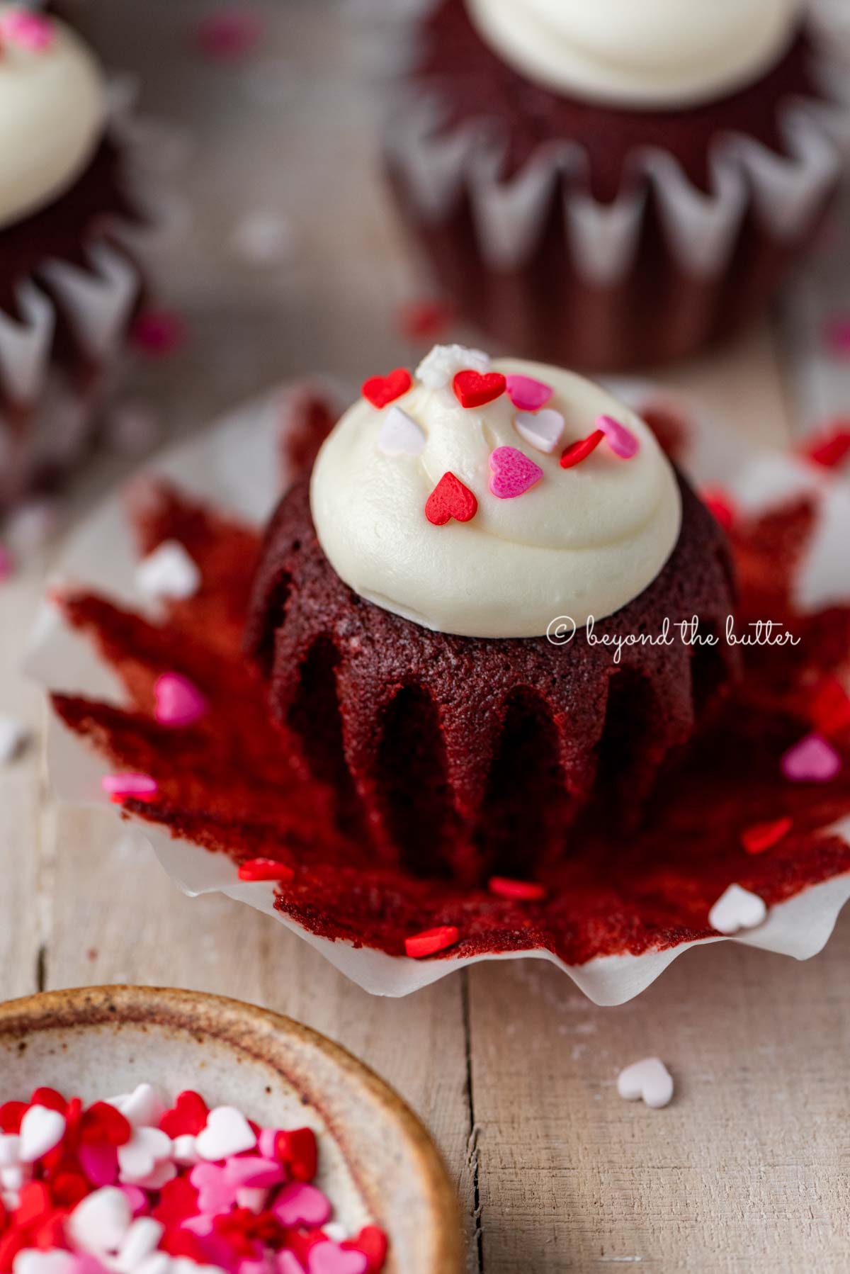 Opened red velvet cupcake with cream cheese frosting topped with heart sprinkles on a light wood background.