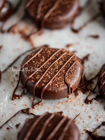 Round peppermint patties on a wax paper lined baking sheet sprinkled with cocoa powder and drizzled with melted chocolate.