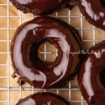 Homemade baked chocolate donuts topped with chocolate ganache glaze on a wire cooling rack.
