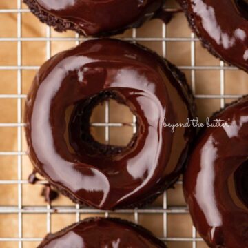Homemade baked chocolate donuts topped with chocolate ganache glaze on a wire cooling rack | © Beyond the Butter®