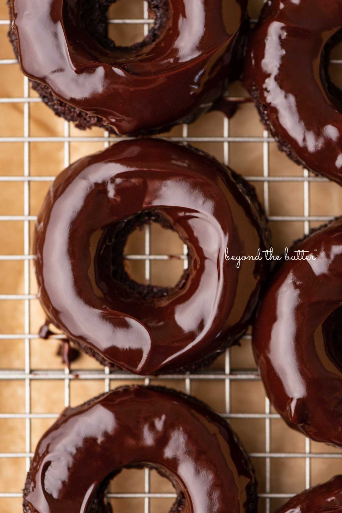 Homemade baked chocolate donuts topped with chocolate ganache glaze on a wire cooling rack.