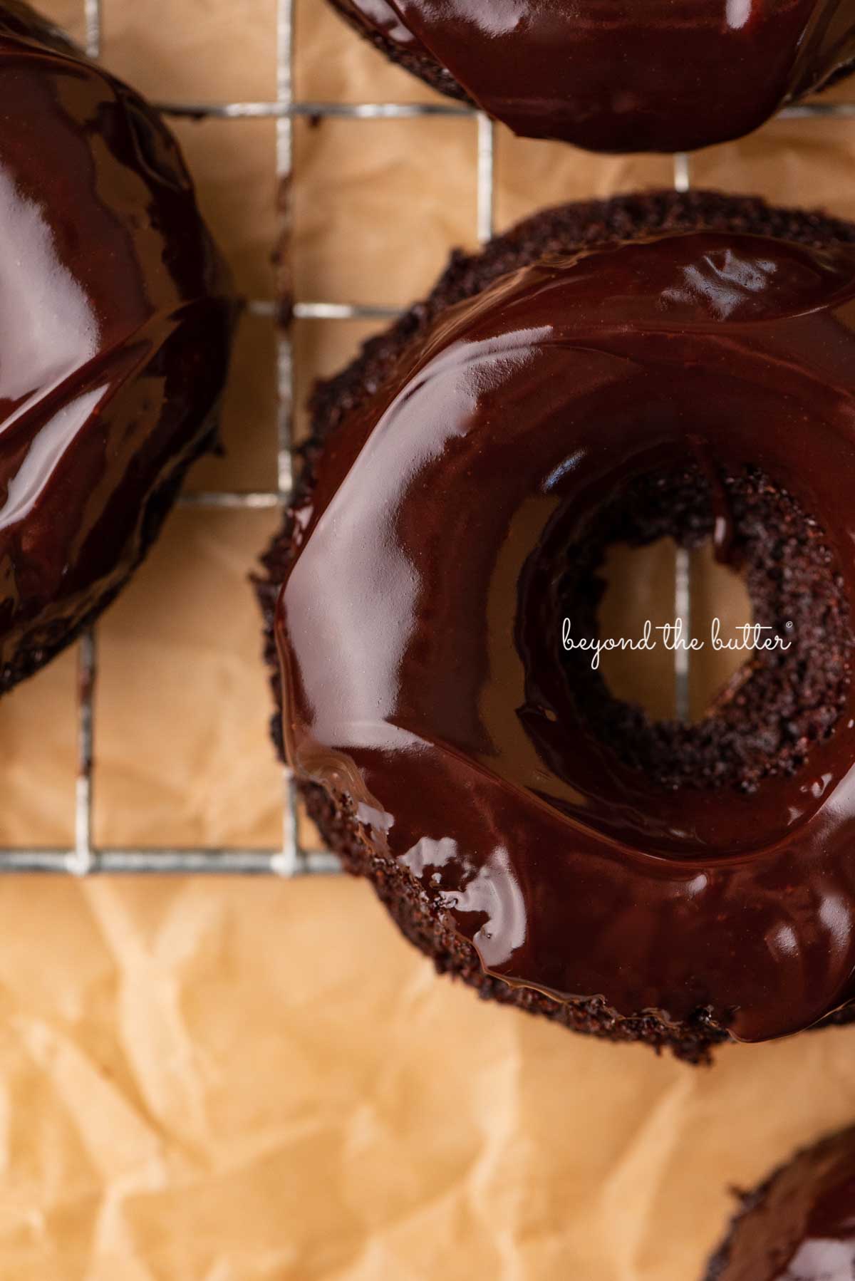 Close up of homemade baked chocolate donuts topped with chocolate ganache on a wire cooling rack | © Beyond the Butter®