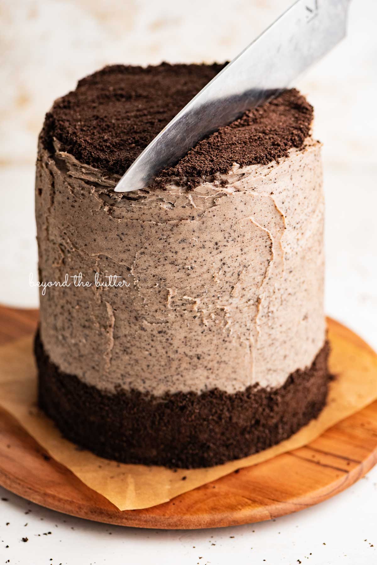 Cutting into chocolate cake with oreo buttercream frosting on parchment paper lined wood cake plate from BeyondtheButter.com | © Beyond the Butter®