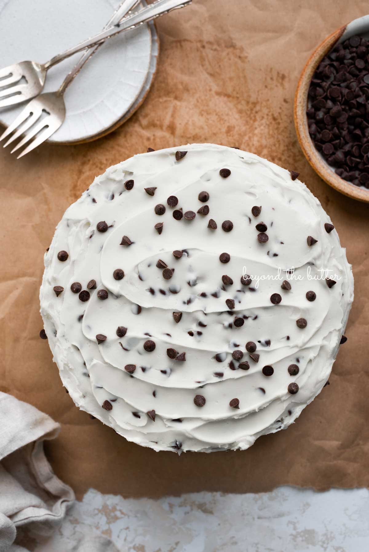 Chocolate chip cake frosted with chocolate chip buttercream frosting with dessert plates, forks, a bowl of chocolate chips, and a napkin on a brown paper background | © Beyond the Butter®