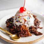 Hot fudge pudding cake topped with ice cream, rainbow jimmies, and a maraschino cherry on a dessert plate and marble white background.