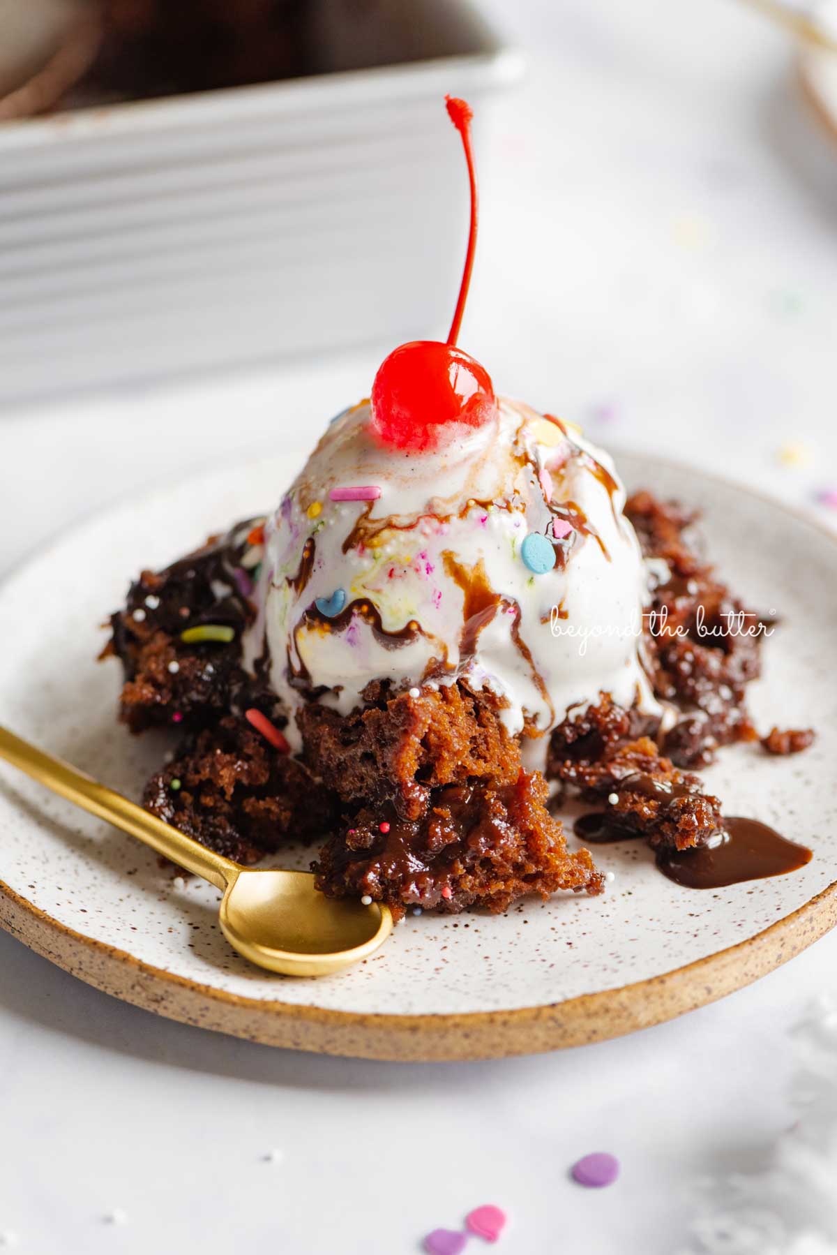 Hot fudge pudding cake topped with ice cream, rainbow jimmies, and a maraschino cherry on a dessert plate and marble white background.