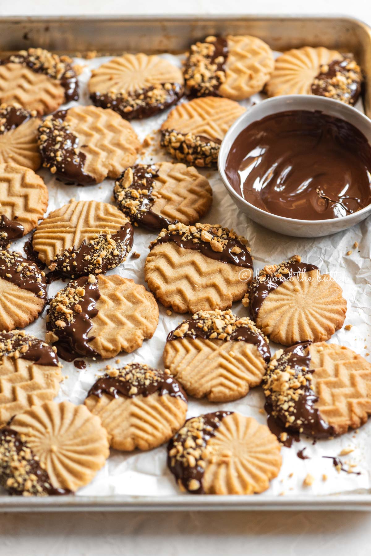 Baking sheet with randomly placed chocolate dipped peanut butter cookies with bowl of chocolate nearby | All Images © Beyond the Butter®