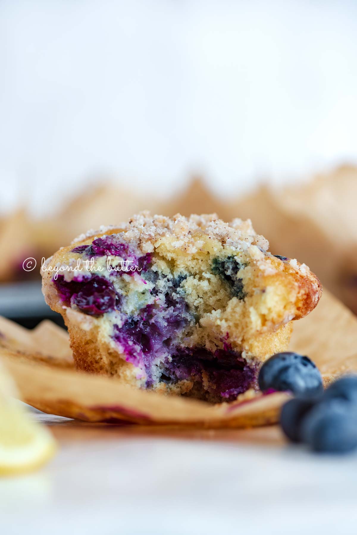 Unwrapped and half eaten lemon blueberry streusel muffin with lemon wedges and blueberries around it | All images © Beyond the Butter®