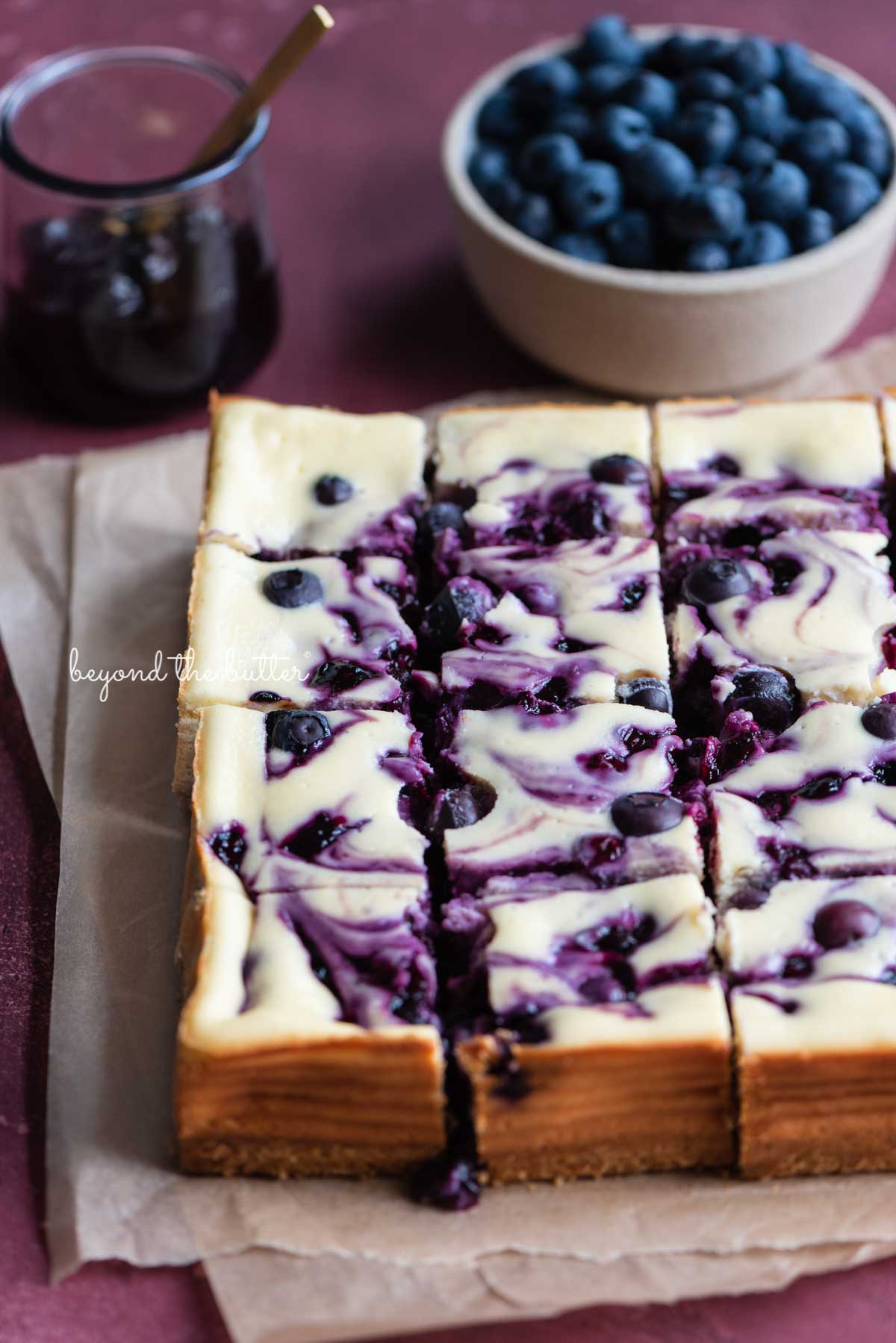 Just sliced homemade blueberry swirl cheesecake bars placed on 2 sheets of parchment paper with bowl of blueberry sauce and fresh blueberries to the side | © Beyond the Butter®