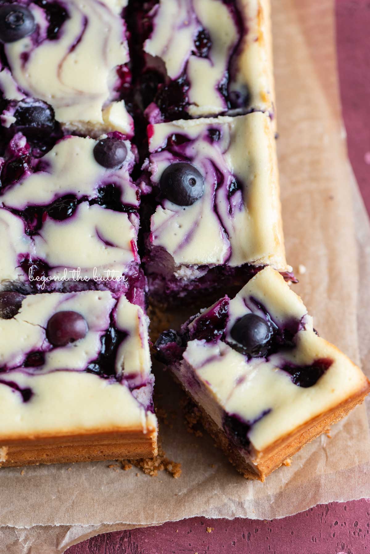 Slices of blueberry swirl cheesecake bar with the corner slice angled on parchment paper | © Beyond the Butter®