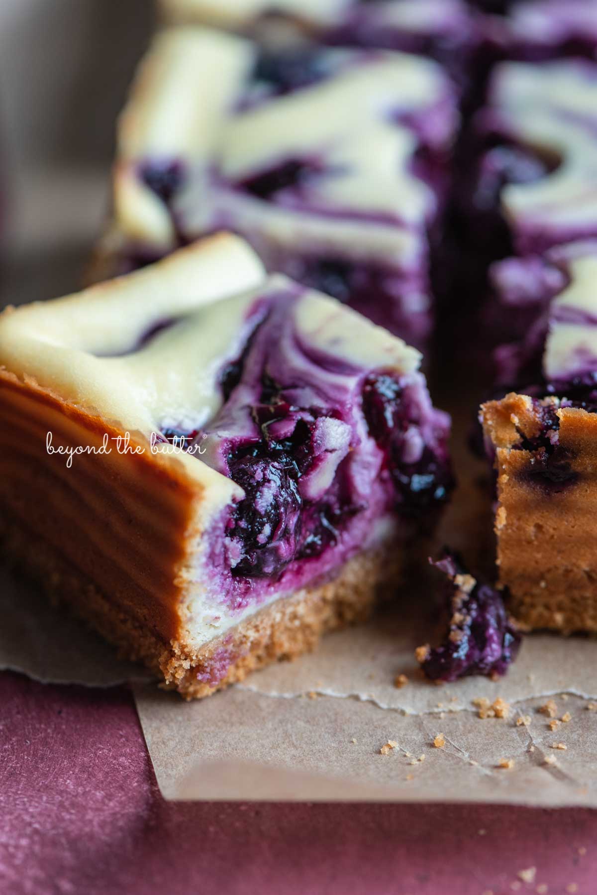 Slice of blueberry swirl cheesecake bar angled to see the side on parchment paper from BeyondtheButter.com | © Beyond the Butter®