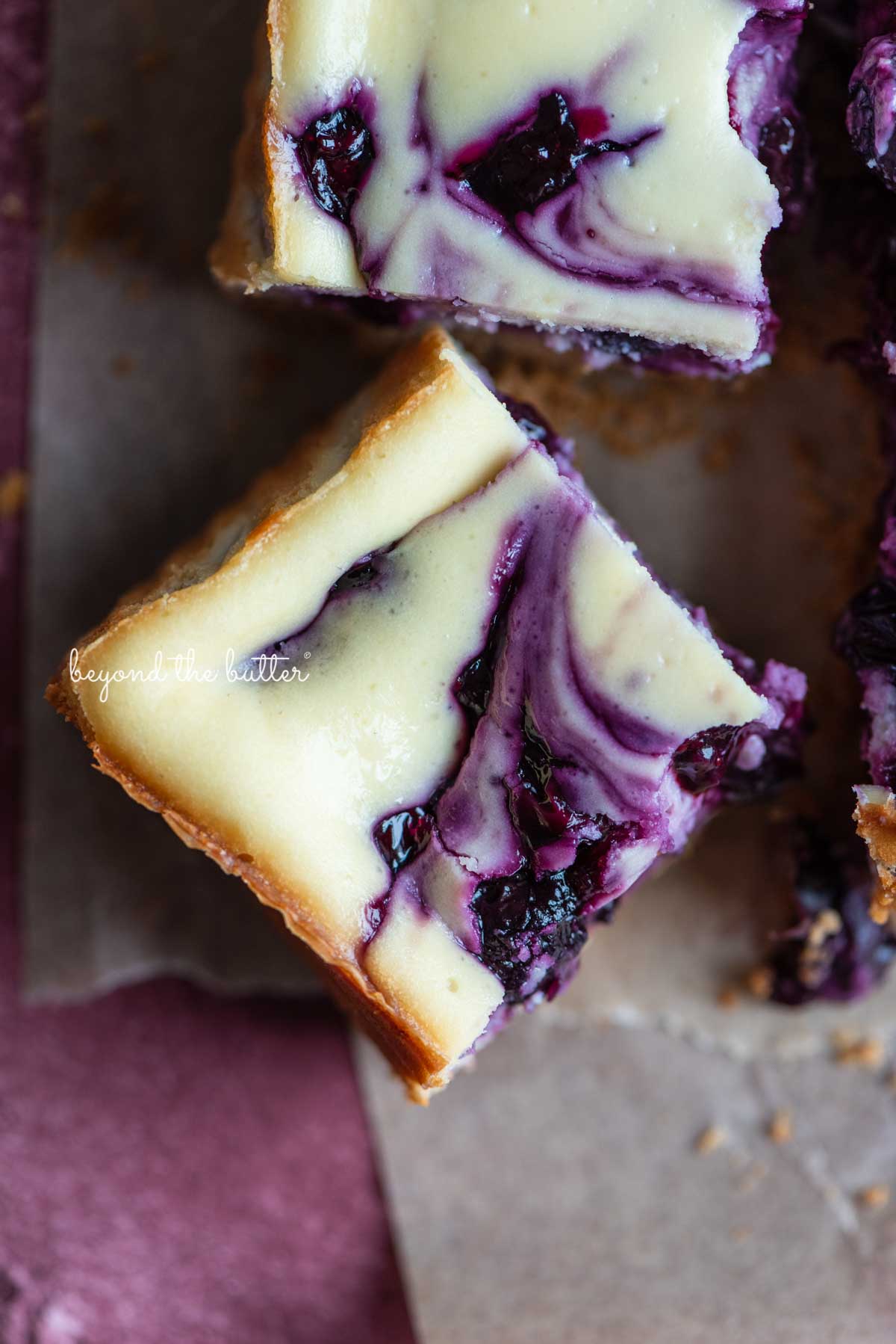Slice of blueberry swirl cheesecake bar angled on parchment paper | © Beyond the Butter®