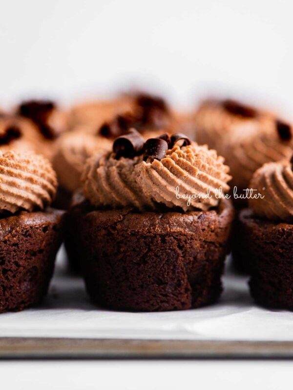 Fudgy brownie bites topped with chocolate mascarpone whipped cream and chocolate curls placed together on a parchment paper lined baking sheet.