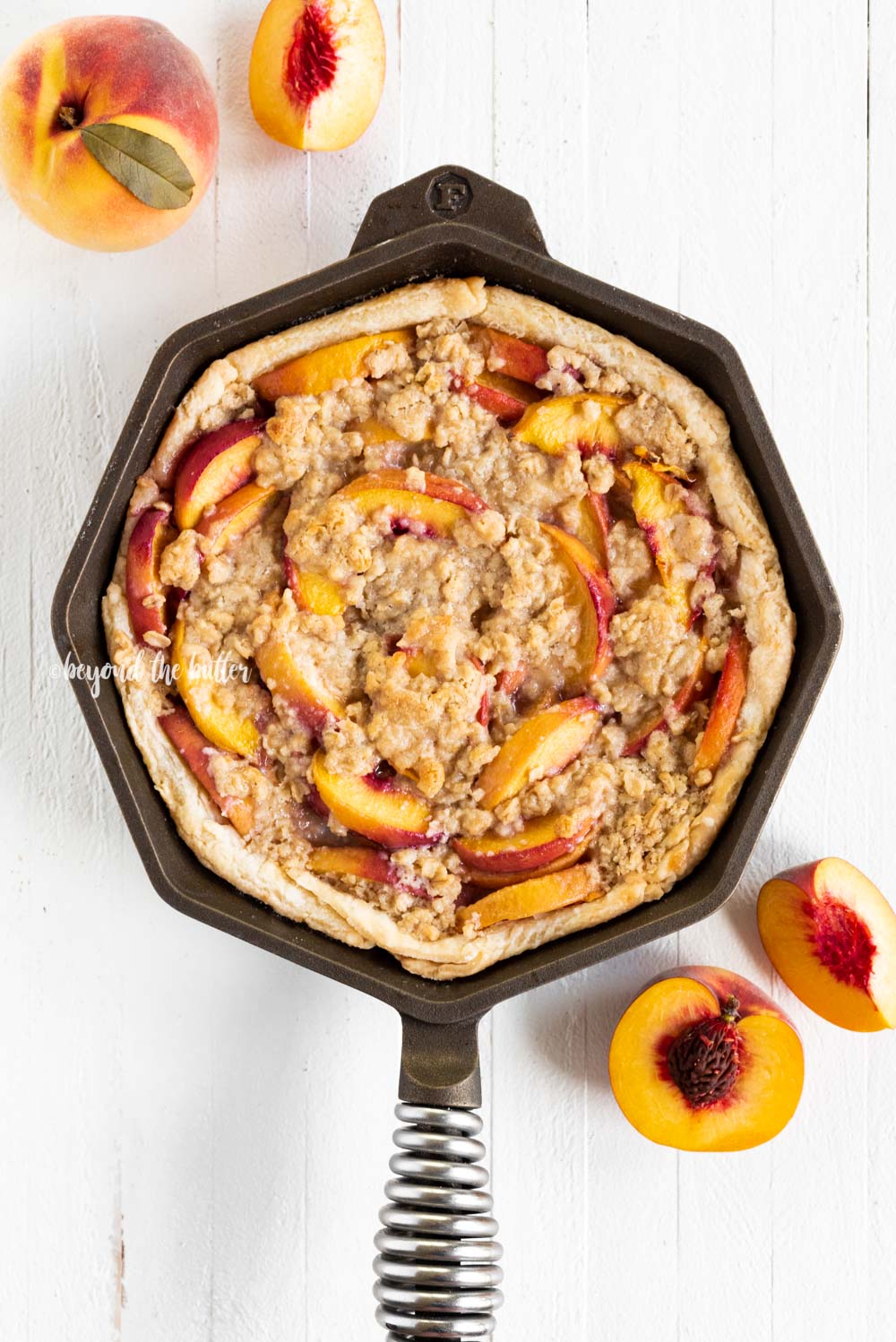 Brown sugar peach crumble pie in cast iron pan with sliced peaches on white wood table.