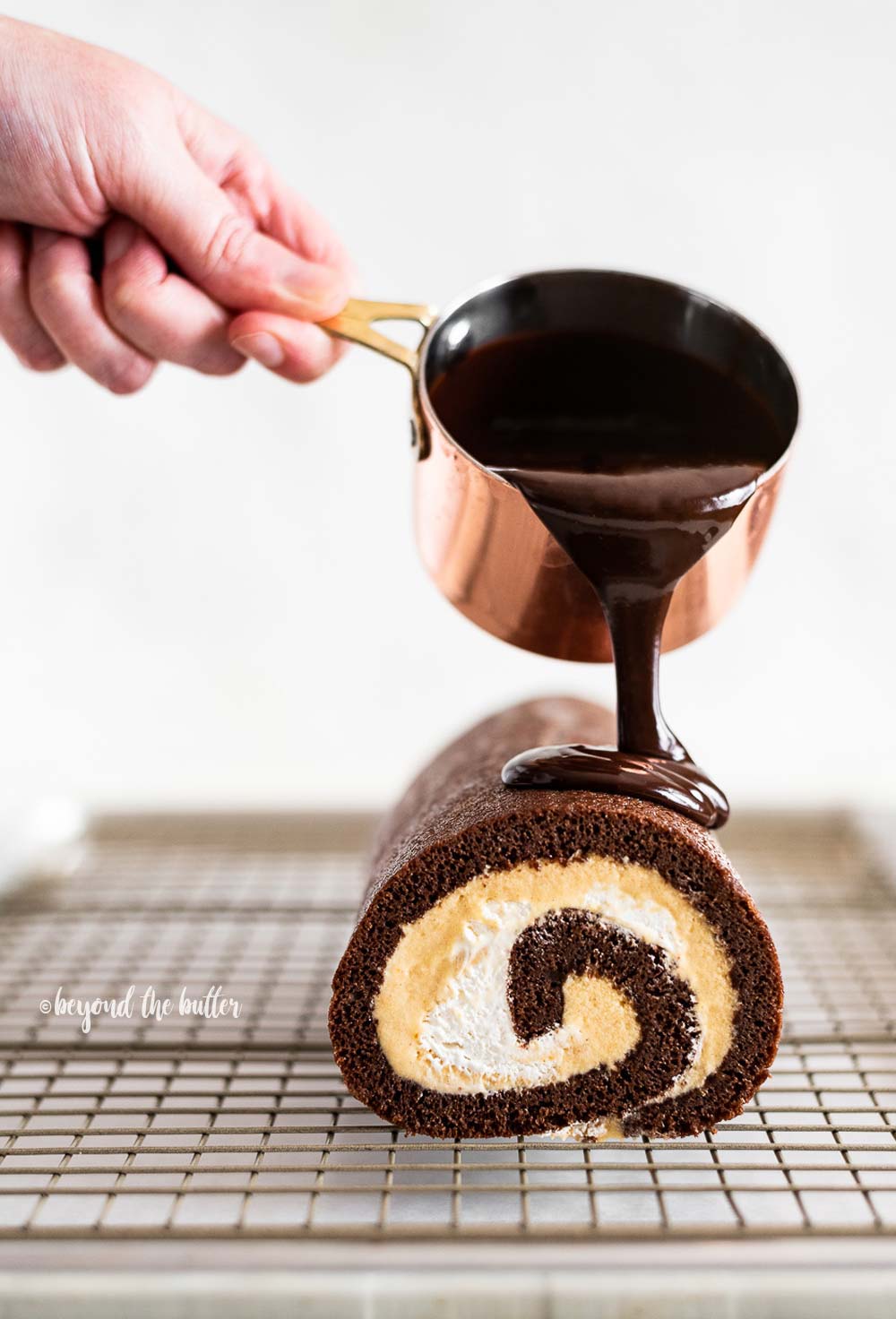 Pouring melted chocolate from small copper pot over chocolate pumpkin roll on wire cooling rack.