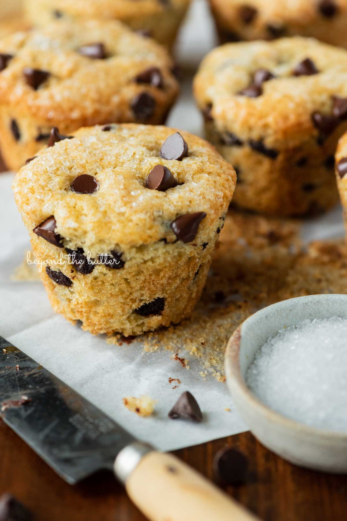 Bakery style chocolate chip mufins unwrapped with a small dipping bowl of sparkling sugar nearby | © Beyond the Butter®
