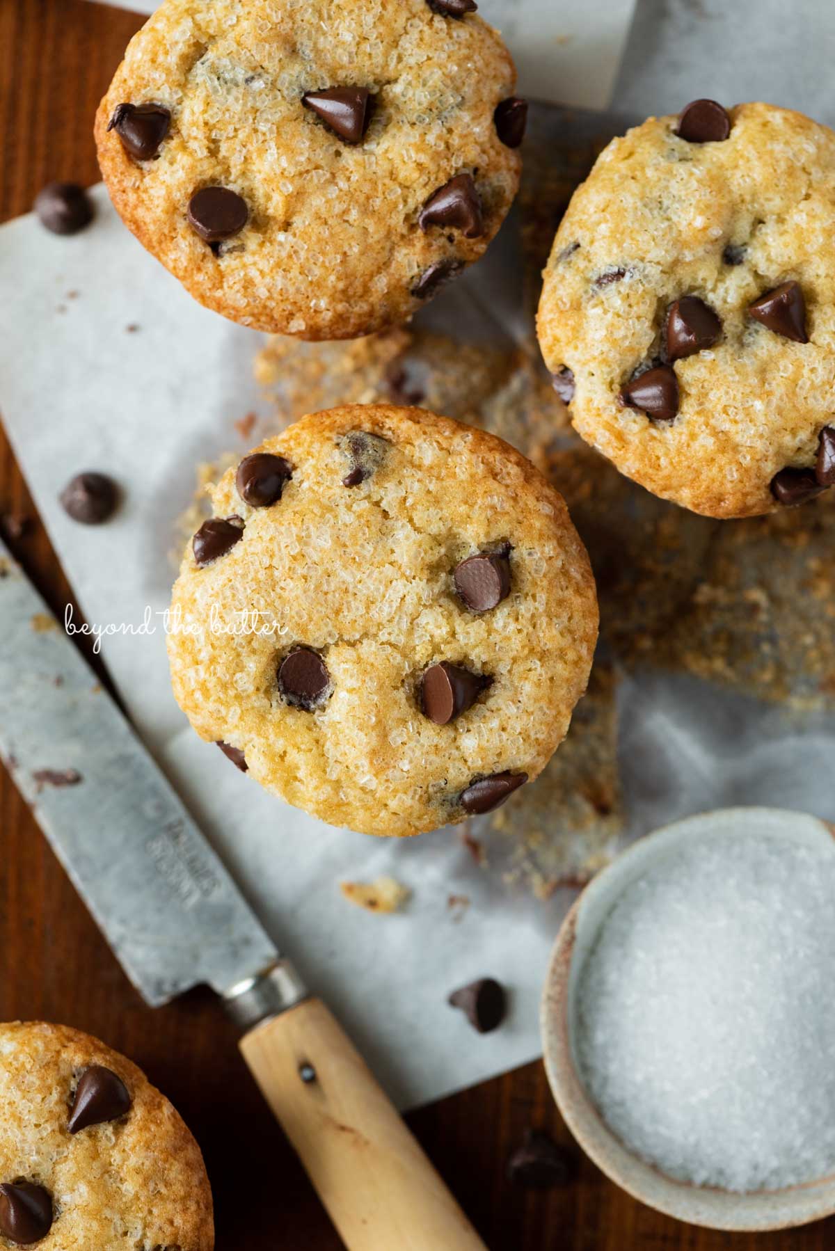 Unwrapped bakery style chocolate chip muffins on flattened muffin liners on a dark background | © Beyond the Butter®