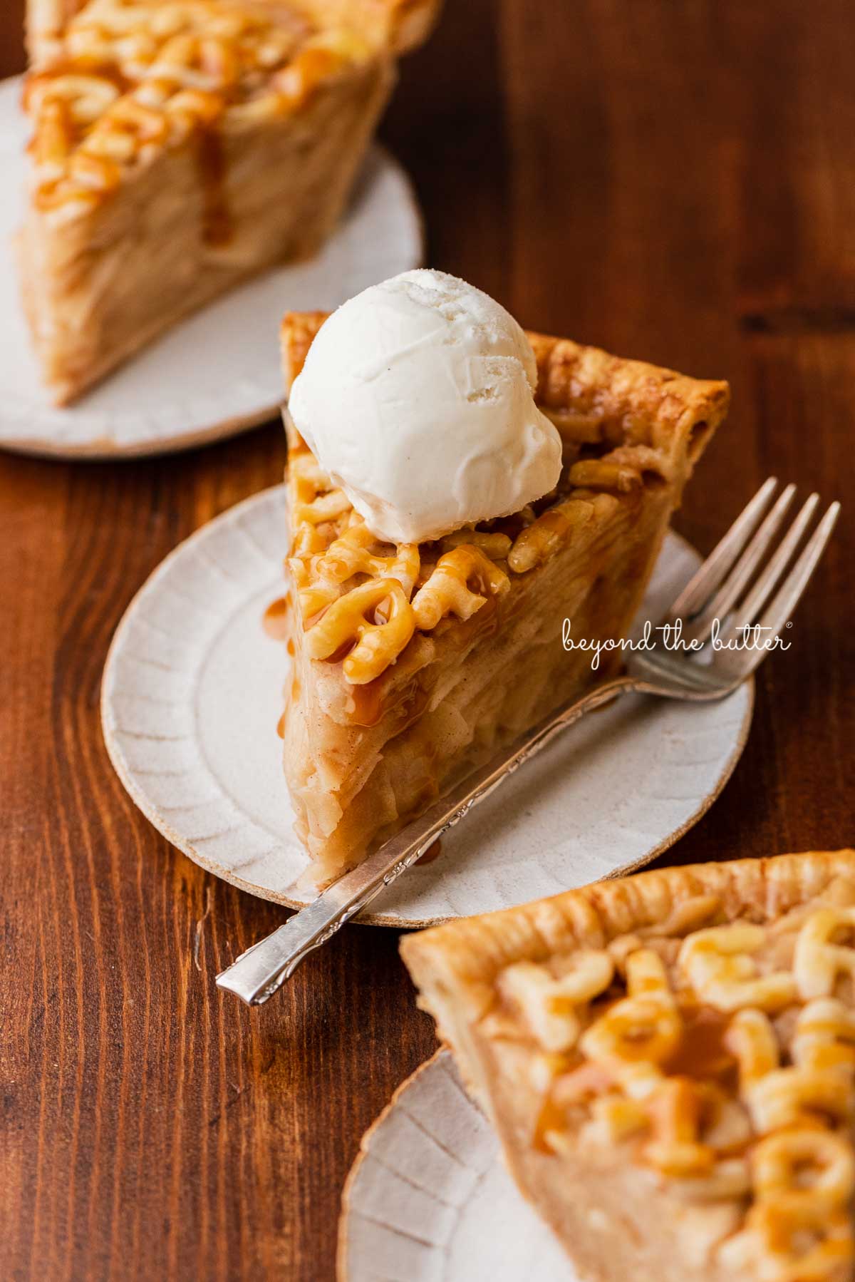Slices of homemade apple pie drizzled with caramel and topped with ice cream on dessert plates and dark wood background | © Beyond the Butter®