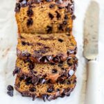 Sliced chocolate chip pumpkin bread with knife and sprinkled chocolate chips on parchment paper lined table.