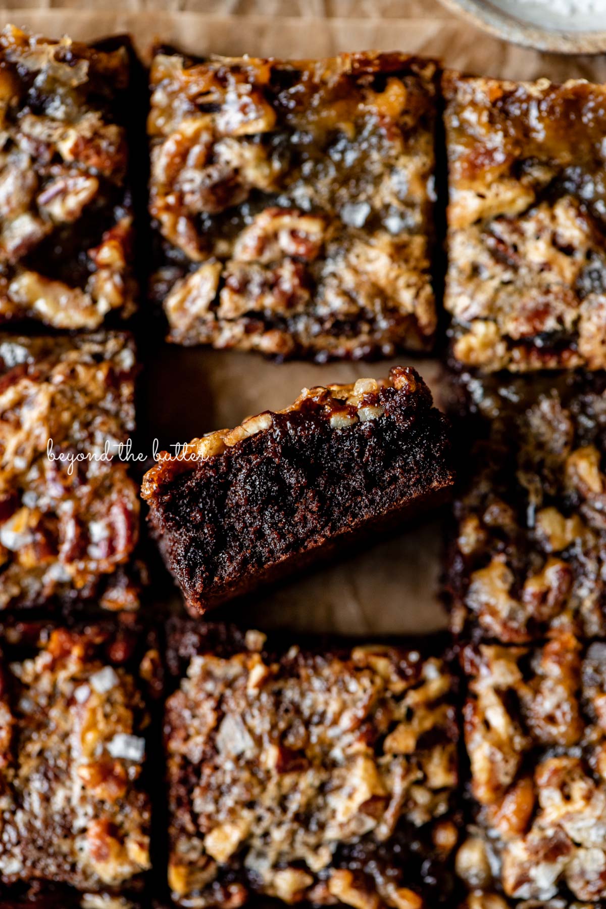 Salted pecan pie brownies placed on brown parchment paper with the center brownie placed on its side | © Beyond the Butter®