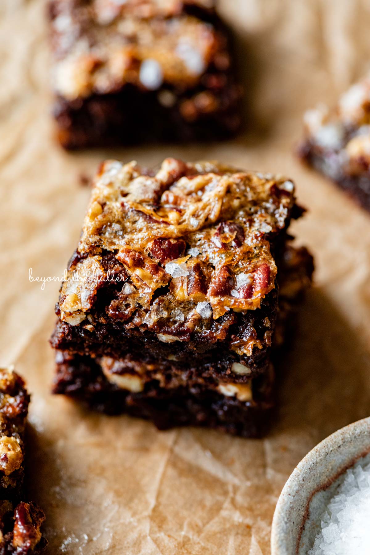 Stacked salted pecan pie brownies on brown parchment paper | © Beyond the Butter®